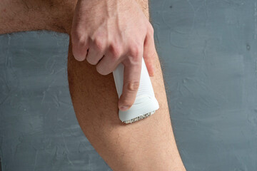 a man shaves his leg hair with a white electric depilator. Men's beauty. - obrazy, fototapety, plakaty