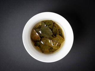 a white bowl with brewed tea leaves on a dark background. Close up, side view. Chinese tea, open leaves. Top view, flat lay