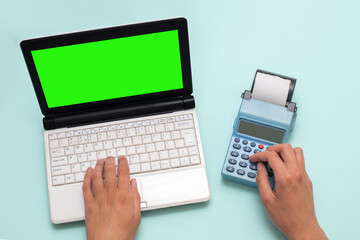 Hand typing on a laptop with a green screen and pressing buttons on a cash register on a blue background