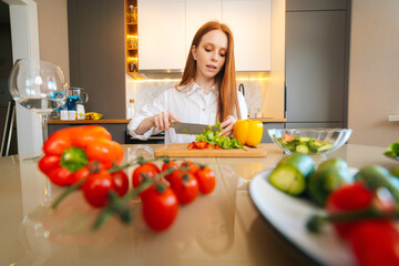 Front low-angle view of young redhead woman cutting fresh lettuce preparing food salad sitting at table in modern kitchen room. Pretty female cooking vegetarian dieting salad full of vitamins.