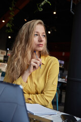 beautiful young caucasian business woman sitting indoors, writing notes in notebook. pretty female sitting at table of cafe, studying with laptop, working. distant work, online education, remote job