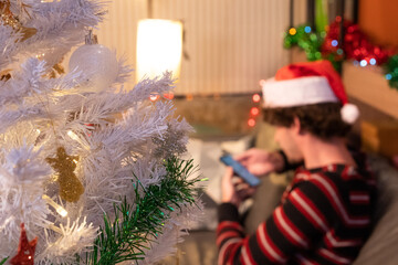 Close-up and focus of a half decorated white Christmas tree on a blurred background of young Caucasian man in a striped sweater wearing a Santa hat sitting using a smartphone on sofa in a living room.