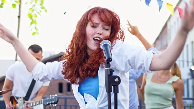 Handsome energetic caucasian woman is singing song on a rooftop party. Band with guitarists, drummer and singer performing at a concert on party.