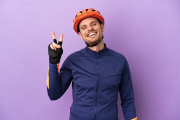 Young Brazilian cyclist man isolated on purple background smiling and showing victory sign