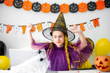 Halloween holiday concept. Cute little girl in witch costume sitting behind a table in Halloween theme decorated room.