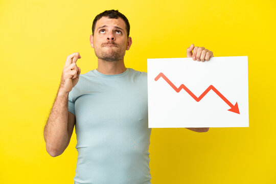 Brazilian man over isolated purple background holding a sign with a decreasing statistics arrow symbol with fingers crossing