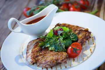 A piece of fried meat lies on a white plate. Decorated with vegetables and herbs. There is a red sauce nearby. The concept of recipes from the restaurant