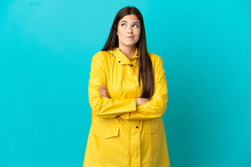 Teenager Brazilian girl wearing a rainproof coat over isolated blue background making doubts...