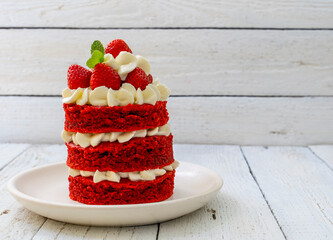 Delicious mini cake red velvet with white cream and strawberries on a saucer on a white wooden background. Food background. Gourmet dessert with berries and a mint leaf.