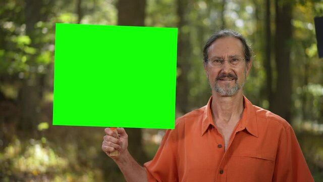 Happy, smiling mature man holding blank green screen chroma key sign in a forest.