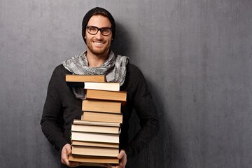 Happy young smart good looking man holding pile of books, smiling leaning against gray wall. Copy space.