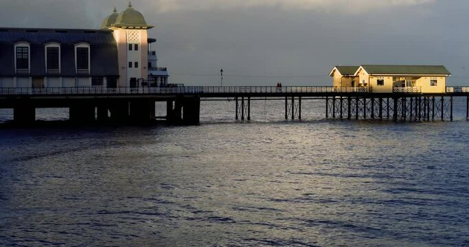 Penarth Pier at high tide: Golden hour at this Welsh tourist attraction and the last rays of setting sunlight hits the pier at dusk. The sea is calm and in the foreground and creates a tranquil scene.