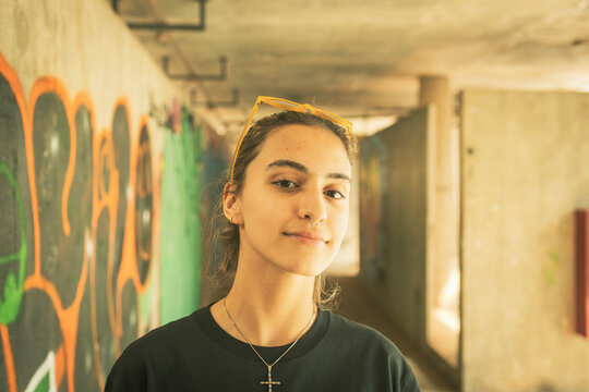 Urban teenager girl wearing long black t-shirt feeling free after covid pandemic staring at the camera in front of a hallway covered by graffiti painting artwork. Youth freedom post pandemic feel.