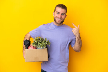 Young caucasian making a move while picking up a box full of things isolated on yellow background...