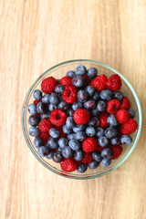 Glass bowl filled with raspberries and blueberries on wooden table. Top view.