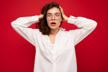 Attractive young emotional shocked amazed curly brunette woman wearing white shirt and optical glasses isolated on red background with copy space