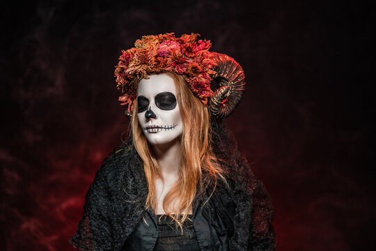 Halloween makeup idea in form of Catrina calavera skeleton, or Faunus with horns, dried flowers. All saints, a witch in rags, with sewn-up mouth, animal skull in a dark red room closed her eyes