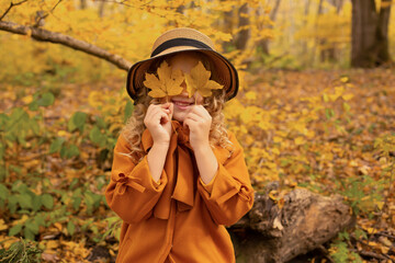 a beautiful blonde girl in a mustard-colored coat and a hat sits on a bench in the autumn forest and covers her eyes with yellow leaves