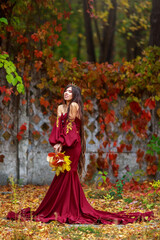 Fototapeta premium Young lady in luxurious long burgundy dress with crown on her head in colorful autumn park