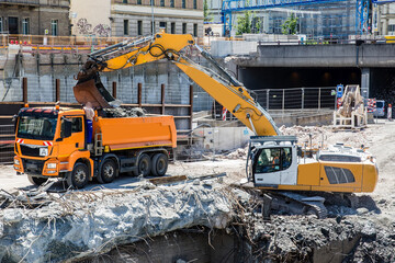 An excavator at a demolition site of a former subway station loads a dump truck.