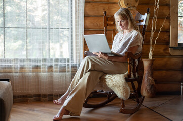 A blonde woman in beige home clothes sits in a rocking chair with a laptop in her hands in a cozy room with large windows and wooden walls. Freelancing and distance learning.