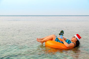 A woman in a swimming ring in a Santa Claus hat with a cocktail and a small Christmas tree in her hand floats in the sea during Christmas holidays.