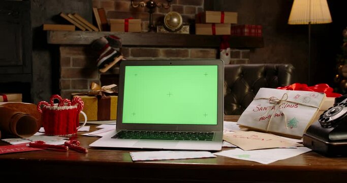 Laptop with green screen at Santa Claus workplace. Close-up of computer with chroma key on table. Christmas time, holidays and celebrations concept. New Year tree and fireplace on background.