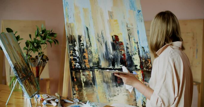 Female artist using palette knife to paint with oil paints on canvas in workshop. Woman painter drawing with oil paints landscape on canvas in art studio
