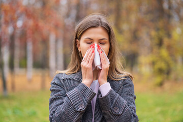A girl stands on the street in a park in autumn and blows her nose in a handkerchief
