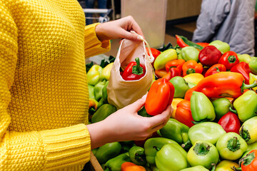 A woman in a yellow sweater puts red and yellow peppers in a cotton fabric ECO bag. The buyer's choice of vegetables in the store. Reusable eco bag for shopping. Zero waste concept.