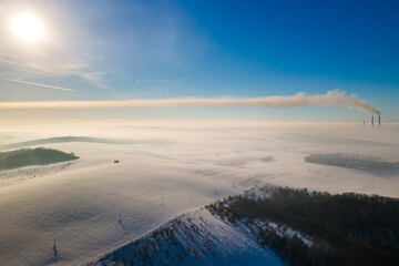 Obraz premium Aerial view of winter landscape with foggy countryside and distant factory pipes emmiting black dirty smoke polluting environment.