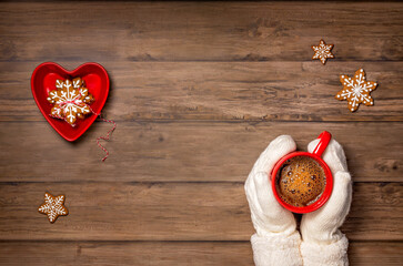 Cozy cup of hot coffee in a red mug held by mittens with christmas cookies.