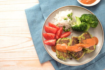 Vegetarian patties of broccoli and flaked oats with tomato, rice, vegetables and spicy sauce, blue napkin and light wooden table, copy space, top view from above