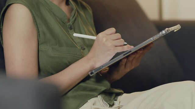 Asian female reading news and trading stock on her digital tablet while sitting on the sofa in the living room home.