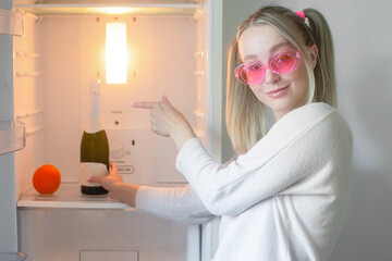 Fototapeta na wymiar A young cheerful girl in pink glasses, opened an empty refrigerator in which there is only a bottle of champagne and an orange and points at them with a finger. Retro style