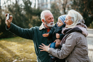 Happy senior couple taking selfie photo with their adorable grandson in public city park.