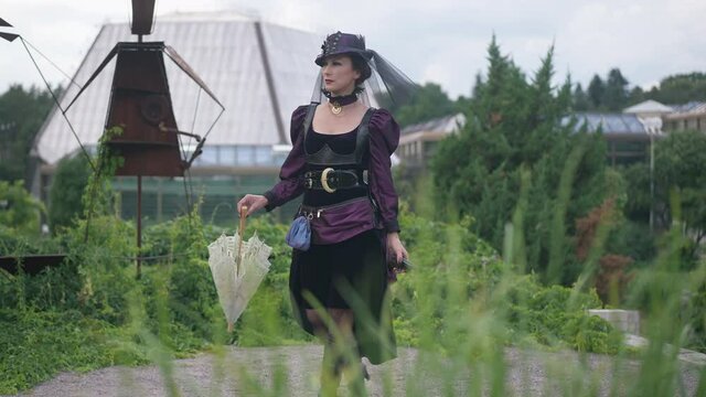 Confident slim woman in purple steampunk dress and hat walking with sun umbrella and gun leaving. Wide shot portrait of confident serious Caucasian lady in Halloween costume strolling in slow motion