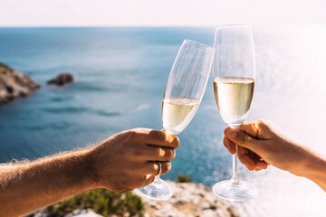Hands holding champagne glasses over the sea. Romantic vacation. Two hands holding champagne glasses on the background of the sea. Toast with champagne glasses on the seashore. Copy space