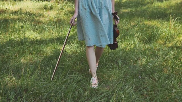 A girl in a dress walks with a violin in a city park.