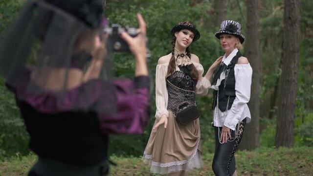 Slim women posing for friend taking photos on camera in forest outdoors. Positive confident Caucasian ladies in Halloween steampunk outfit enjoying holiday celebration in woods. Slow motion