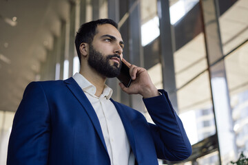 Portrait of handsome pensive middle eastern businessman talking on mobile phone looking at window standing in modern office