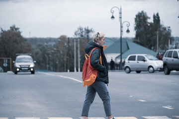 a girl with a backpack crosses the road on the carriageway at a pedestrian crossing in front of passing cars - obrazy, fototapety, plakaty