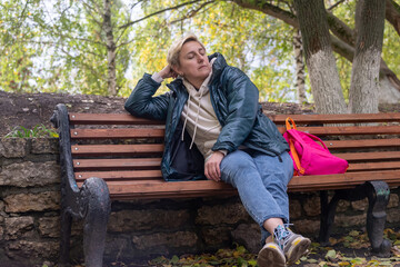 Fototapeta premium a girl in a dark jacket and with an orange backpack is sitting on a bench against the background of autumn nature, thinking and looking ahead