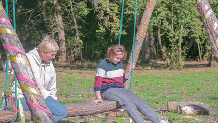 mom and daughter are riding on a long wooden swing in nature in the bright sun