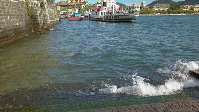 Small waves hitting a concrete pier in Lake Como, in the city of Como, Lombardy, Italy. In the background there is a tourist ferry, and a good view to the beautiful city of Como. 