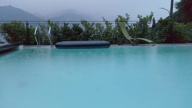 Rain drops falling into a swimming pool, Lake Como, Lombardy, Italy. It is an overcast, rainy summer day. There is an air mattress and sun chair behind the pool. In the background there are mountains