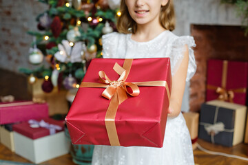 Fototapeta na wymiar Selective focus. A girl in a white dress with a red gift box close-up in a room decorated with a Christmas tree and a fireplace.