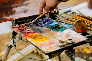 Shallow depth of field (selective focus) image with a painter and his color palette while painting a landscape on a sunny autumn day.