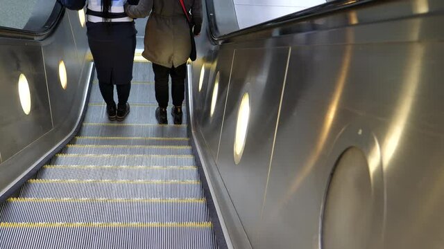 Rear view of two unidentified women going down by escalator at subway or underground station. Blind woman with cane is holding onto sighted guide as they reaching end of escalator.