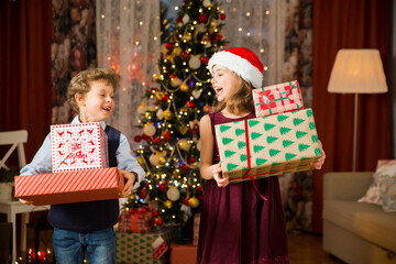 Obraz na płótnie Canvas Excited cute kids happily smiling, playing with a pile of Christmas gifts. Beautifully decorated living room with lights and Christmas tree. Children having fun, celebrating family holiday
